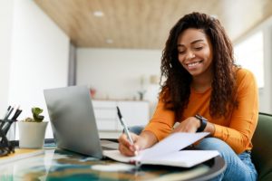 smiling woman sitting at desk in front of laptop writing in notebook studying for Arizona Insurance License Exam